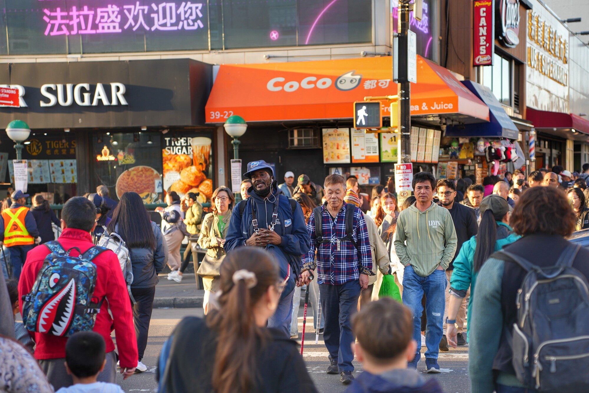 Three pedestrians cross beneath an LED screen reading “Welcome to Flushing.” Queens, Nov. 8, 2025.