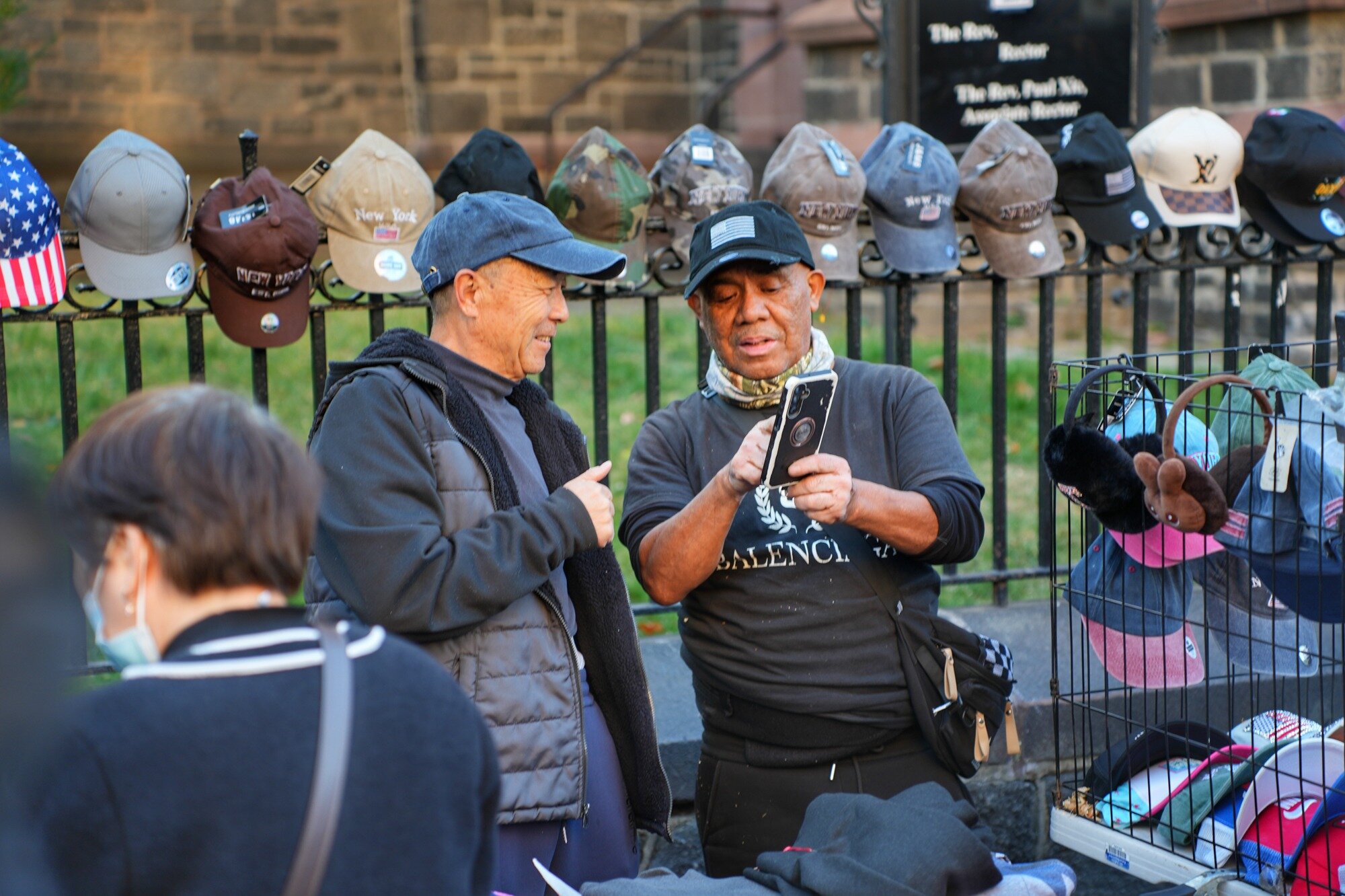 Two street vendors look at a phone beside a hat display outside a church. Nov. 8, 2025.